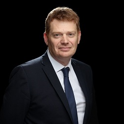 Man in a dark suit dark tie white shirt against a dark background