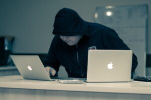 Man in dark clothing with hood up stooped over two mac laptops suggesting criminal intent