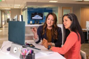 Two women sitting at a desk in business attire discussing something on a computer screen