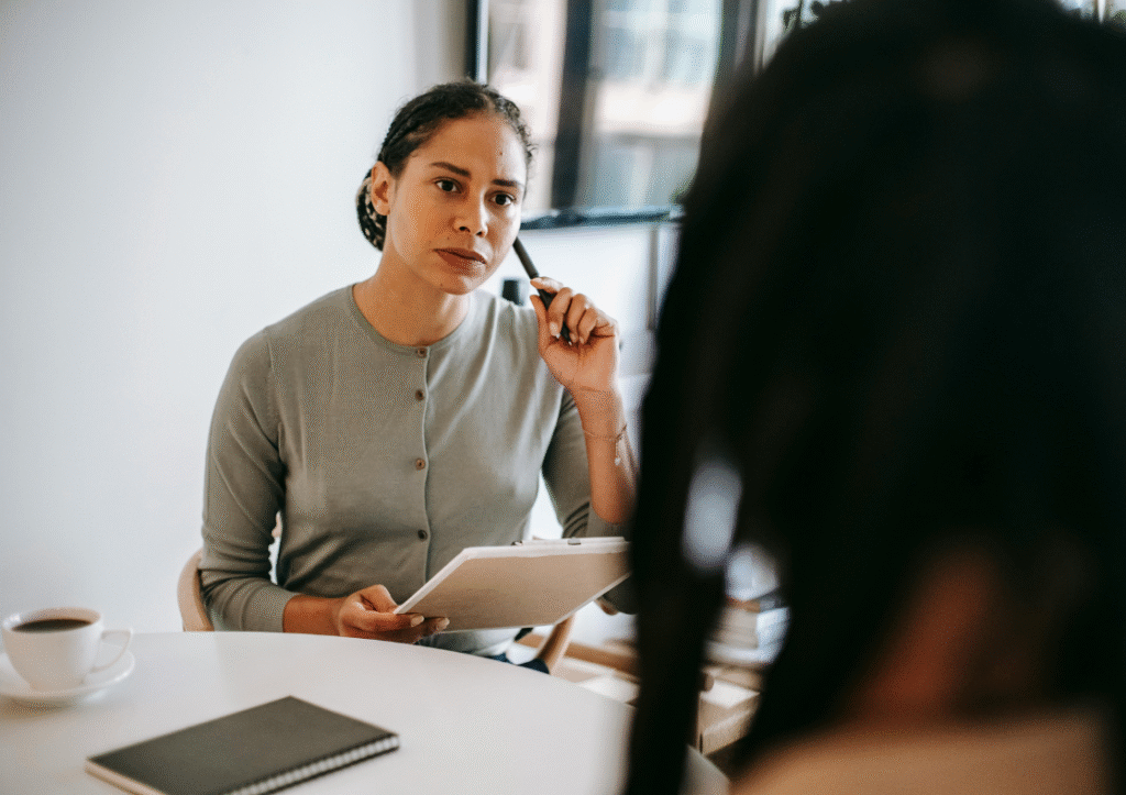 Two women in an office setting have a conversation, one with a serious look on her face, making meeting notes