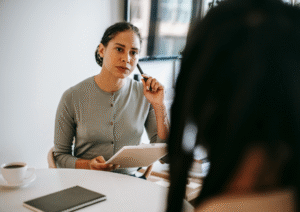 Two women in an office setting have a conversation, one with a serious look on her face, making meeting notes
