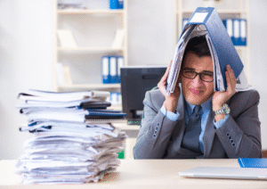 Business man sitting at a desk with a stack of paperwork in front of him holding a folder over his head as if to try and hide from it