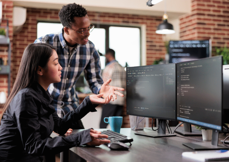 Two colleagues in an office setting looking at two monitors depicting coding having a conversation about it