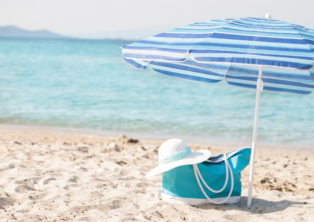 A sunny beach scene with a blue beach bag, white summer hat, and blue and white stripy parasol
