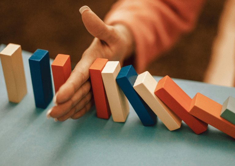 Hand stopping colourful wooden blocks from falling in a domino-style effect. Blocks are orange, blue and cream. To reflect SRA sectoral risk assessment.