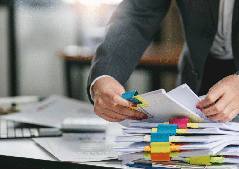 pile of paperwork on a desk with brightly coloured bulldog clips. A pair of female hands sorting the paperwork in an office