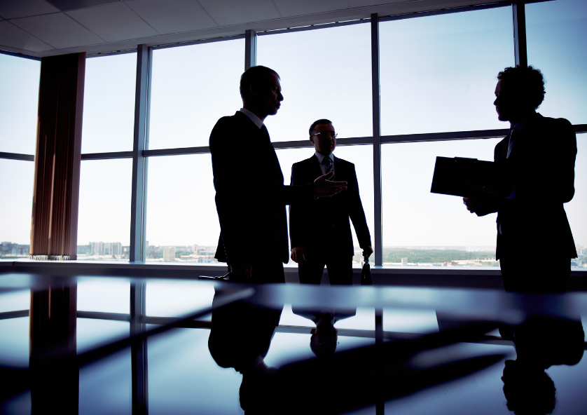 Shadowy figures standing aroud a glass office boardroom table,